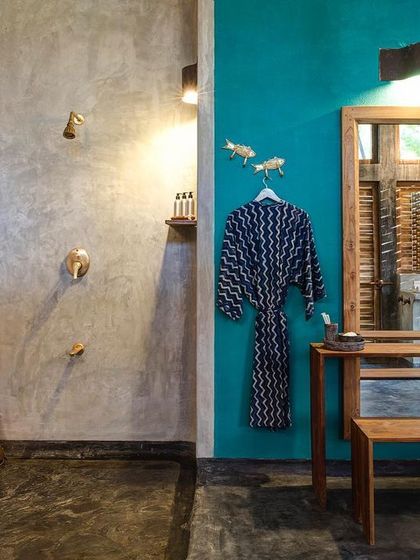 The bathroom of the private villa combines a vibrant turquoise accent wall with raw cement textures and custom wooden furniture. The open shower area enhances the feeling of tropical living.