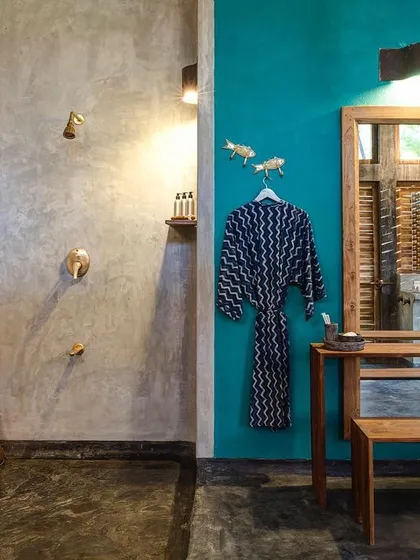 The bathroom of the private villa combines a vibrant turquoise accent wall with raw cement textures and custom wooden furniture. The open shower area enhances the feeling of tropical living.