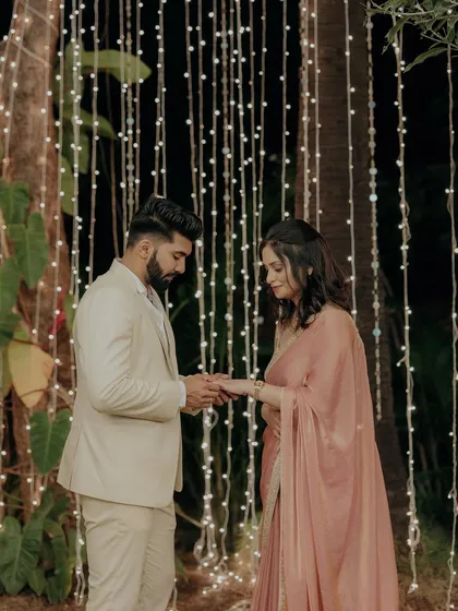 The ring exchange moment during an intimate engagement ceremony, set against a curtain of warm fairy lights.