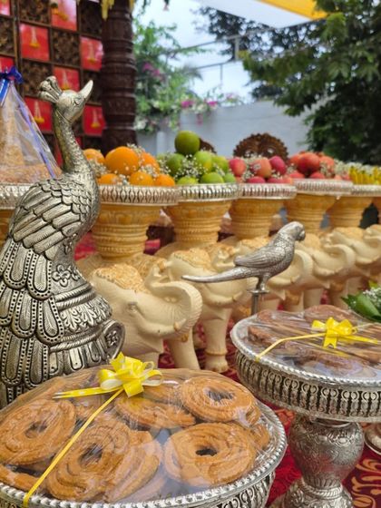 A close-up of traditional sweets like murukku (chakli) and fruits displayed in silver bowls on elephant-themed stands.