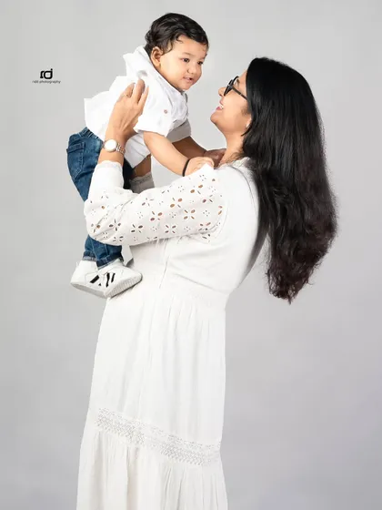 A mother lifts her son, and they share a beautiful, smiling gaze. The simple grey background and white outfits keep the focus on their loving interaction.
