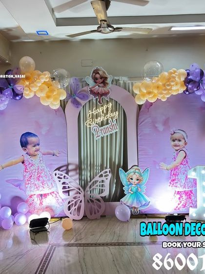 An elaborate fairy theme for a first birthday, featuring a three-panel backdrop with custom photos. The scene is decorated with purple and yellow balloons, illuminated butterfly wings, and multiple fairy cutouts.
