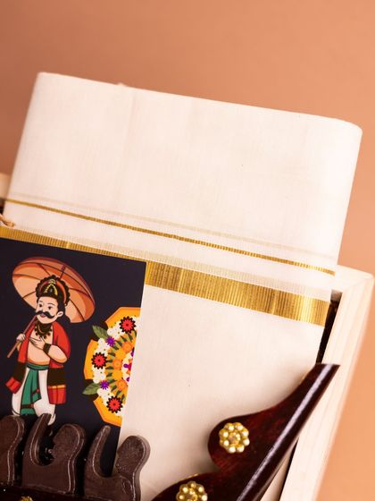 A detailed shot of the 'Onasanchari' hamper, focusing on the crisp, gold-bordered kasavu mundu and the charming Mahabali greeting card. These elements are central to an authentic Onam celebration.