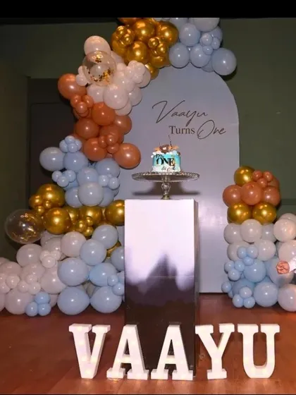A stylish first birthday for Vaayu. This modern setup uses a muted color palette of blue, brown, and gold balloons against an arched backdrop, with large letters spelling out the name.