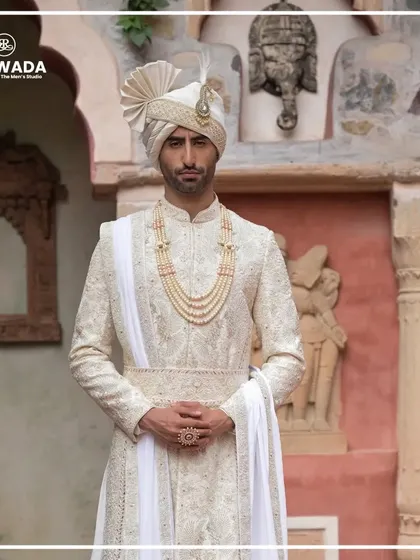 A full-length view of the groom against a heritage backdrop. The white-on-white embroidery and the flowing silhouette create a look of pure, timeless elegance.