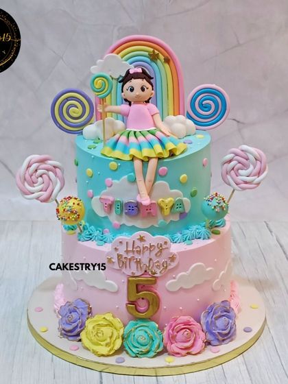 A magical two-tier Candyland rainbow cake for a fifth birthday. This black forest cake features a doll topper, colorful lollipops, and multi-colored roses against a pastel rainbow background.