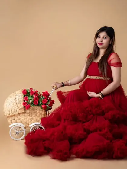 A lovely studio portrait featuring a mom-to-be in a dramatic red tulle gown, seated next to a charming wicker pram filled with red roses. This setup adds a touch of classic romance to the maternity session.