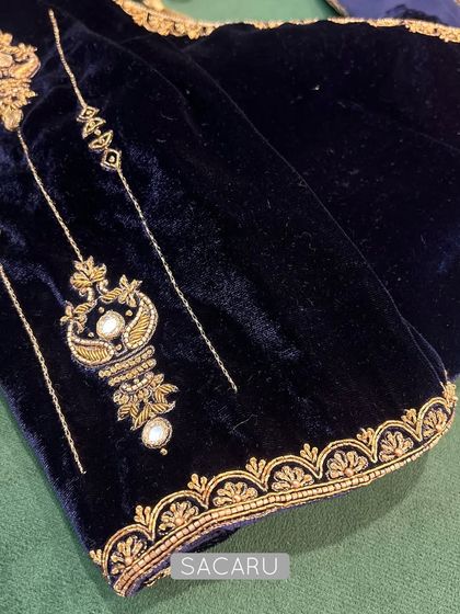 A close-up of a sapphire blue velvet blouse sleeve, showcasing timeless gold embroidery with pearl accents in a classic motif.