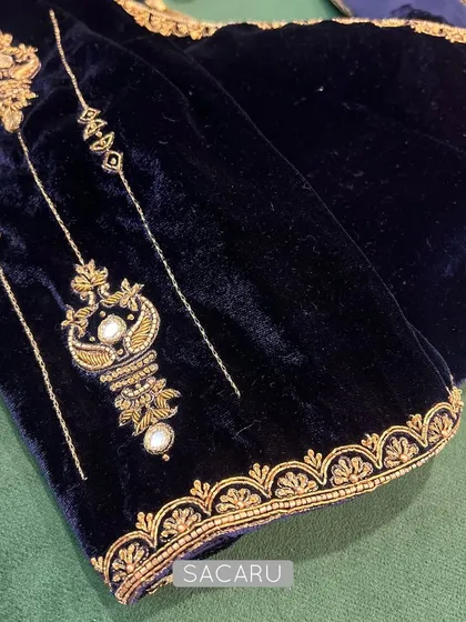 A close-up of a sapphire blue velvet blouse sleeve, showcasing timeless gold embroidery with pearl accents in a classic motif.