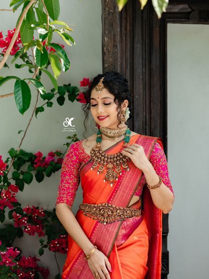 A beautiful bride in a vibrant orange and pink Kanjivaram saree. The makeup is kept fresh and radiant to complement the bright colors of her outfit and antique jewelry.