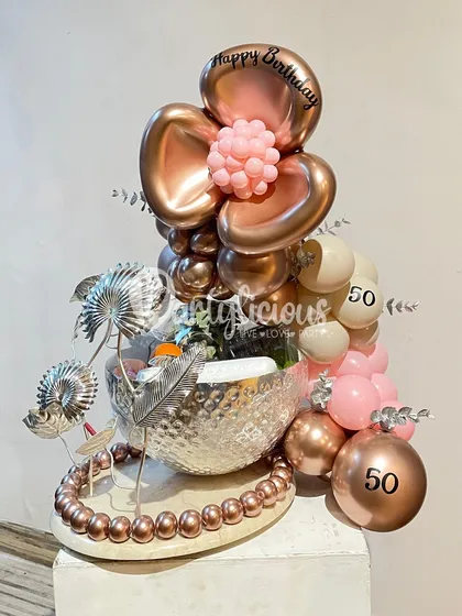 A sophisticated and modern 50th birthday gift arrangement. This piece combines a metallic bowl, silver-plumed accents, and a rose-gold floral balloon bouquet for a truly unique and artistic look.