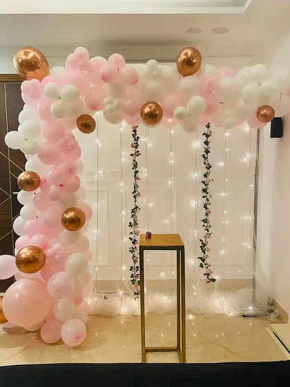An elegant corner decoration with a pink, white, and rose gold balloon arch. The backdrop of fairy lights on a sheer curtain adds a soft, magical glow.