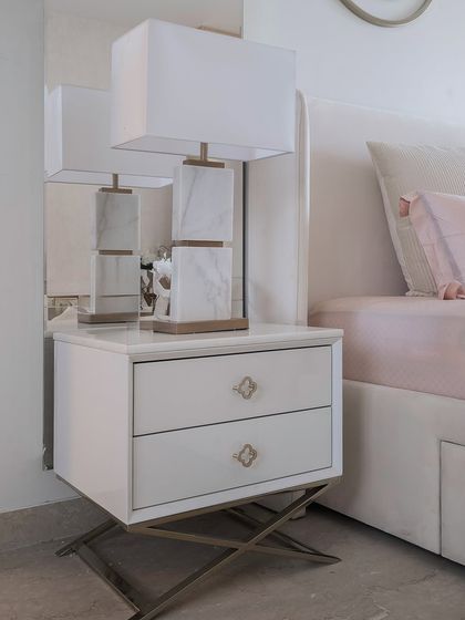 A detail of the sleek white bedside table with high-gloss gold accents and X-shaped legs. It is paired with a sculptural marble lamp that doubles as a piece of art.