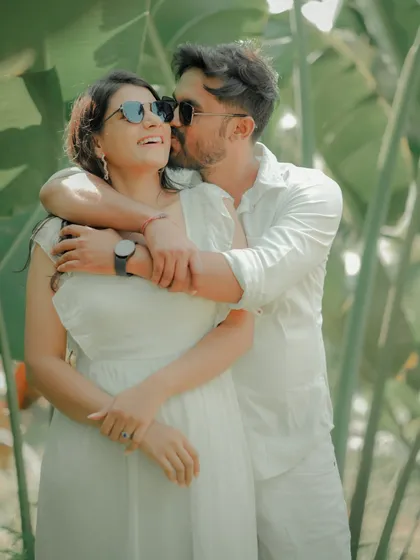 A fun and carefree moment captured against a backdrop of lush tropical leaves. The couple, dressed in matching white, shares a laugh, showcasing the relaxed and joyful side of their relationship.