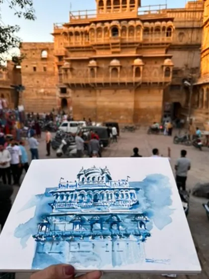 A monochromatic blue watercolour painting of a beautiful haveli in Jaisalmer, held up against the golden sandstone of the actual location.