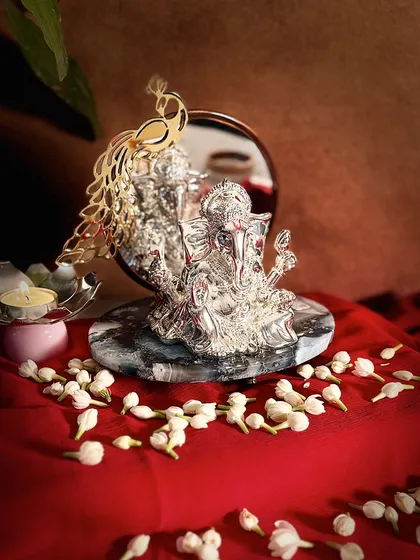 This stunning Ganesha idol combines divine artistry with reflective beauty. The silver-plated form is set against a mirror backdrop adorned with a golden peacock, all resting on a sophisticated grey marble base.