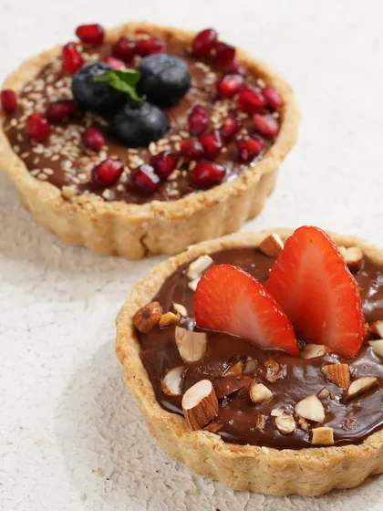 A pair of delicious-looking chocolate tarts, one topped with strawberries and almonds, the other with blueberries and pomegranate. The shot highlights the fresh, quality ingredients.