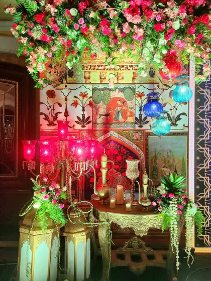 A close-up of the intricate decor for the Sufi Night. This setup includes a backdrop of Mughal paintings, a gilded table with traditional ewers, and colorful glass lanterns, all under a canopy of flowers.