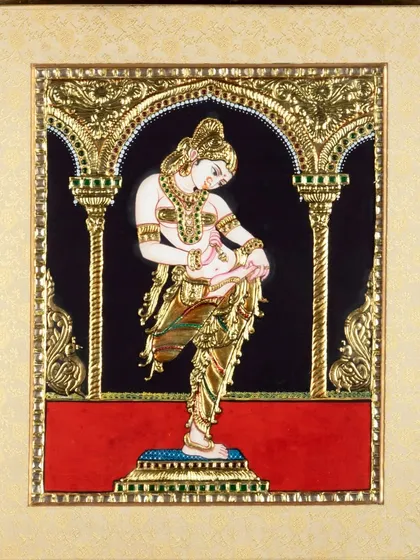 A framed Tanjore painting of a dancer, shot to highlight the intricate gold work and vibrant colors against the black background.