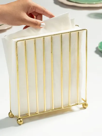 A styled shot of a gold napkin holder, shown in use on a set table. The human element of a hand reaching for a napkin makes the scene feel more dynamic and relatable.