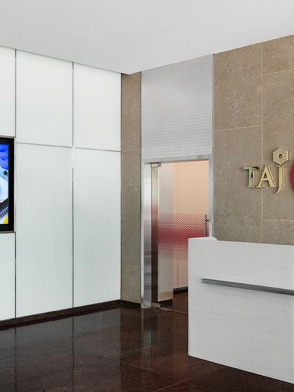 The informal reception area at the top of the stairs in 'The High-Flying Office'. A clean, modern reception desk is set against a stone wall with the TajSATS logo, creating a professional and welcoming first point of contact.