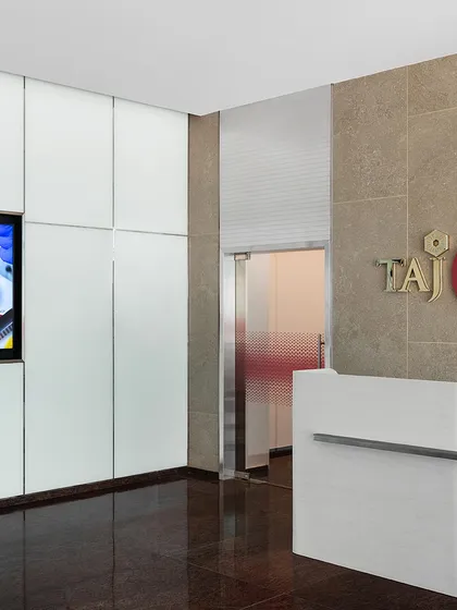 The informal reception area at the top of the stairs in 'The High-Flying Office'. A clean, modern reception desk is set against a stone wall with the TajSATS logo, creating a professional and welcoming first point of contact.