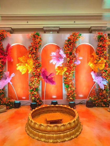 The main stage for the Haldi ceremony, featuring a central brass vessel for the rituals. The backdrop is a warm orange with oversized, colorful paper flowers, creating a modern and artistic take on traditional decor.