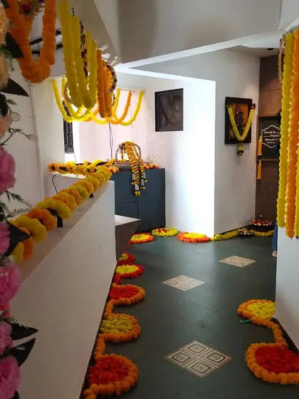 This image shows a hallway decorated for a traditional home ceremony. The floor is lined with rangoli patterns made from marigold petals, and garlands of yellow and orange flowers hang from the ceiling and walls.