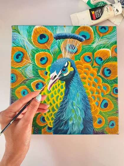 A top-down view of the peacock painting in progress, with my hand and brush for scale. You can see the tubes of paint I use to achieve these vibrant blues, greens, and yellows.