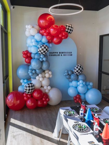 A race car themed balloon garland in red, white, and blue with checkered flag balloons. The custom backdrop wishes Charlie and George a happy birthday.