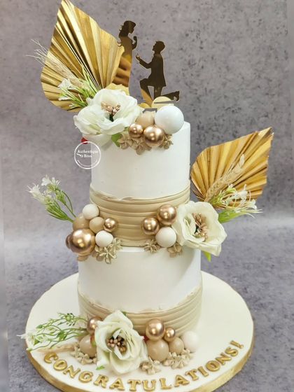 A beautiful boho-themed engagement cake. This two-tiered creation is adorned with dried palm leaves, neutral-toned spheres, and delicate white flowers, complete with a silhouette topper of the proposal moment.