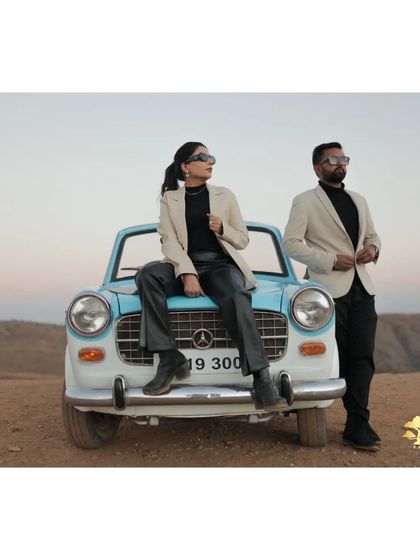 A high-fashion shot with a vintage car in a desert-like landscape. The couple's coordinated modern outfits create a striking contrast with the classic car.