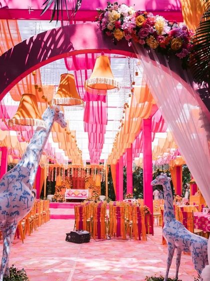 A whimsical entrance to a mehendi ceremony. The archway is flanked by two large, decorated giraffe statues, leading into a space filled with pink and orange drapes.