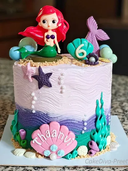 A mer-mazing double-barrel chocolate buttercream cake for a little mermaid fan. This 10-inch tall creation features vibrant under-the-sea colors, handcrafted shells, and a fun Ariel topper, perfect for a magical kids' birthday party.