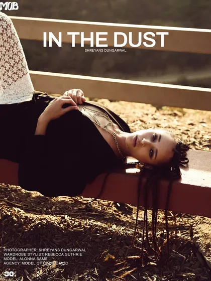 An outdoor editorial for MOB Journal with a rustic, sun-drenched feel. The model's relaxed pose on a wooden fence highlights the layered gold necklaces against a simple black blazer, creating a story of effortless style.