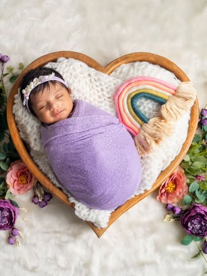 Wrapped in a lovely lavender swaddle, this newborn sleeps peacefully in a heart-shaped bowl, adorned with a rainbow and fresh flowers.