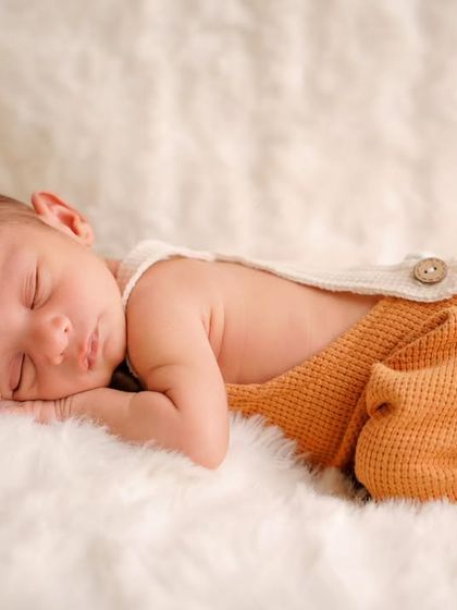 A sleeping newborn wearing cute orange pants lies on their tummy on a fluffy white blanket, a perfect example of a simple baby portrait.