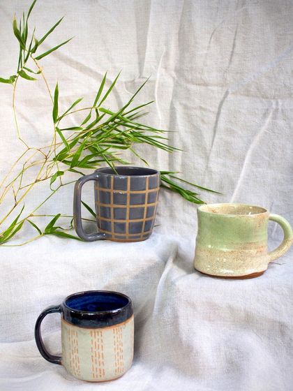 Another view of the three student-made mugs, set against a backdrop of bamboo leaves.