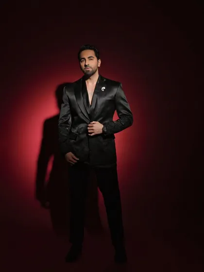 A full-length portrait of Ayushmann Khurrana for a Pinkvilla style feature. The deep red background and satin suit create a look of pure sophistication.