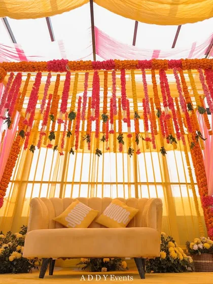 A bright and cheerful Haldi stage, decorated with drapes of yellow and pink fabric and cascading strings of marigolds.