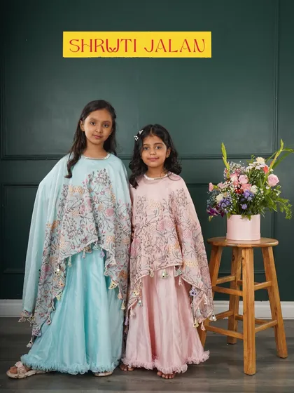 Two sisters ready to twirl in their matching cape-style gowns. The flowing capes feature a lovely floral print and playful tassels, one in sky blue and the other in blush pink.