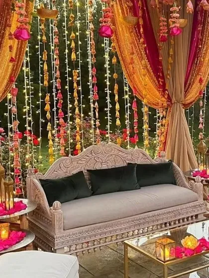 An intimate and cozy seating arrangement for a Sangeet or Mehendi, with a carved sofa, rich fabrics, and warm lighting from lanterns and fairy lights.