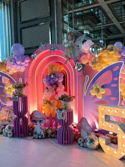 A vibrant and magical fairy-themed 5th birthday party. The decor is filled with color, featuring rainbow-arched backdrops, large butterfly wings, mushroom props, and a light-up number "5".