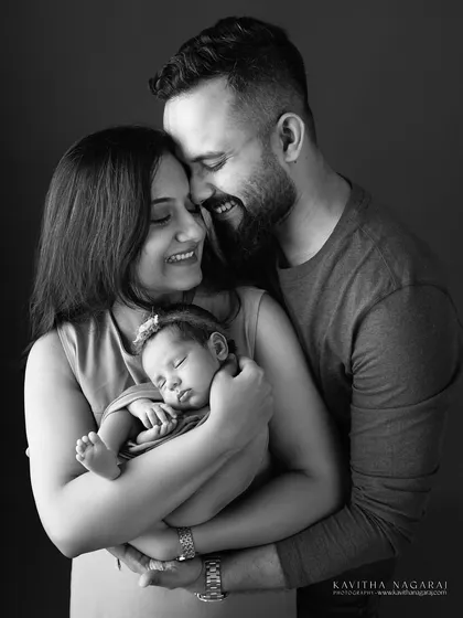 A black and white version of a classic family portrait. The focus is on the emotional connection between the parents and their sleeping newborn, a timeless memory.