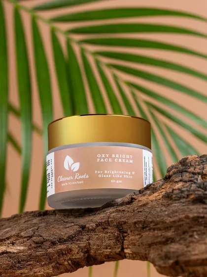 A shot of Cleaner Roots' Oxy Bright Face Cream, styled with a palm leaf and tree bark. The natural props and lighting create a warm, tropical, and organic vibe.