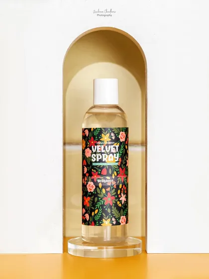 A clean, elegant shot of a body spray framed within a white arch prop. The simplicity of the composition puts all the focus on the product's vibrant label.