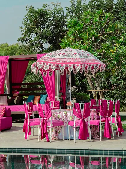 A poolside dining setup for a bhaat ceremony. The all-pink decor, including a cabana, draped chairs, and a printed parasol, creates a chic and celebratory atmosphere.