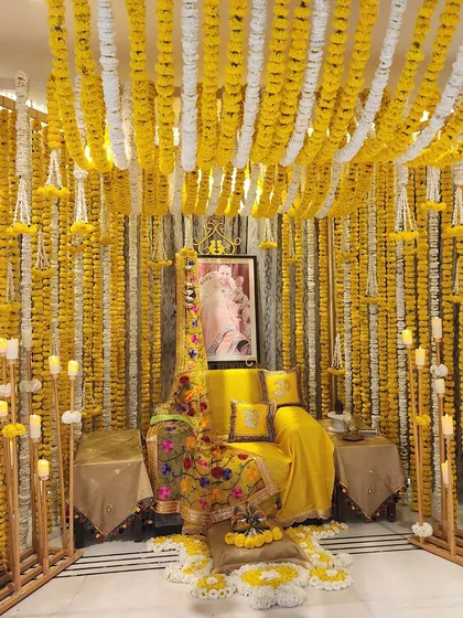 A stunning setup for a Satsang evening at home, bathed in auspicious yellow. The space is enveloped in curtains of yellow and white marigolds, with a comfortable seating area and the warm glow of many candles.