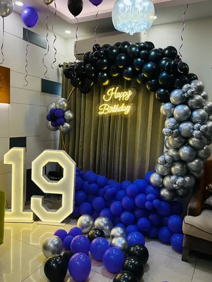 I can match any theme, including this cool and stylish 19th birthday party. The decoration features a moody color palette of purple, black, and silver balloons, a 'Happy Birthday' neon sign, and a large light-up marquee number '19'.