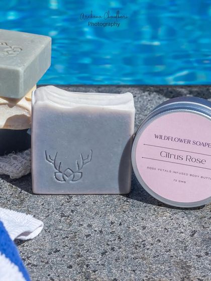 A detailed shot focusing on one bar of soap and the body butter tin. The bright, direct sunlight highlights the textures of the soap and the poolside stone.